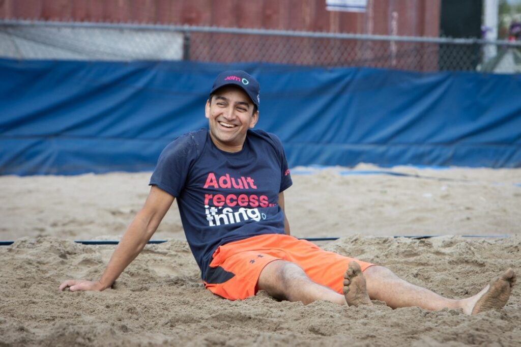 Sandeep Khembavi, Manager, Fincnail Planning & Analytics in Finance at JAM. Sandeep's sitting in the on a beach volleyball court wearing a JAM hat and t-shirt that says "Adult recess... It's a thing."