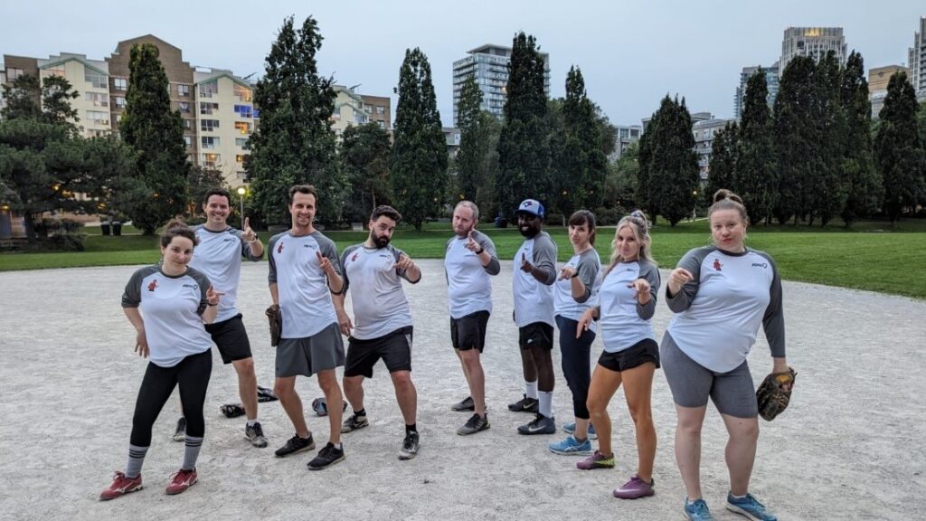 JAM corporate softball team. They are wearing white tshirts with the JAM logo and popular "You just got JAMmed" meme from the TV show, Parks & Rec. The team is posing with a hip popped out and a finger point, imitating the meme